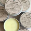 Foot-Balm-Open round yellow foot balm tin, open, surrounded by closed tins