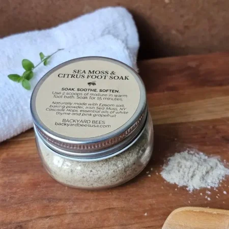 Glass jar of Sea Moss & Citrus Foot Soak on a wooden board with a wooden scoop, white towel, spilled soak, and fresh green sprig — natural Bee Barefoot product photo.