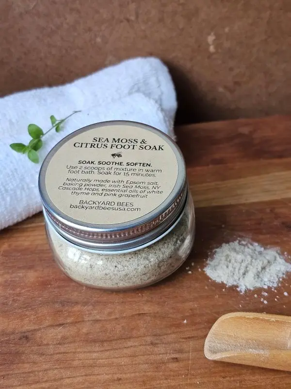 Glass jar of Sea Moss & Citrus Foot Soak on a wooden board with a wooden scoop, white towel, spilled soak, and fresh green sprig — natural Bee Barefoot product photo.