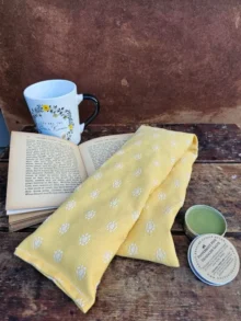 Heatable lavender neck pillow with flaxseed and lavender, shown with open book, mug and headache salve for cozy relaxation