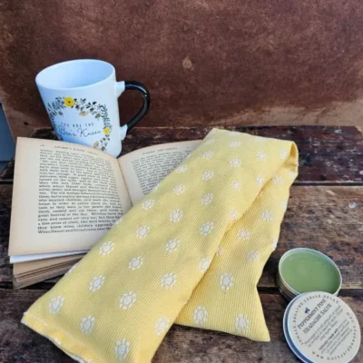 Heatable lavender neck pillow with flaxseed and lavender, shown with open book, mug and headache salve for cozy relaxation