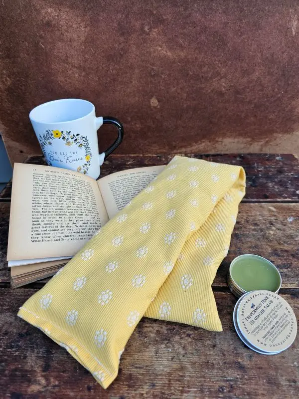 Heatable lavender neck pillow with flaxseed and lavender, shown with open book, mug and headache salve for cozy relaxation