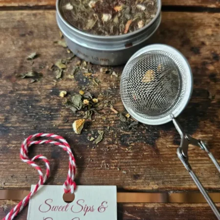 Holiday Take Tea And See Closeup Loose-leaf tea tin beside a filled mesh tea strainer and Sweet Sips & Cozy Wishes tag on a rustic wooden surface.