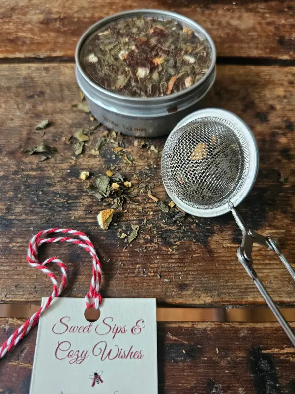 Holiday Take Tea And See Closeup Loose-leaf tea tin beside a filled mesh tea strainer and Sweet Sips & Cozy Wishes tag on a rustic wooden surface.