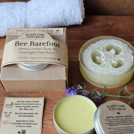 Bee Barefoot Duo set with Honey Loofah Foot Soap, Sleepy Time Foot Balm, white towel, and greenery on a wooden board — calm natural flat lay