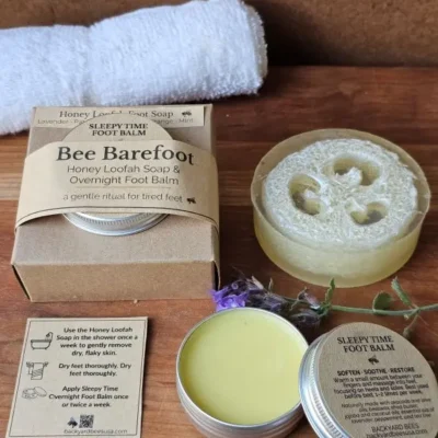 Open tin of Sleepy Time Foot Balm beside Honey Loofah Soap, greenery, and ritual card on warm wood — soothing Bee Barefoot detail image.