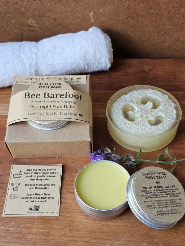 Open tin of Sleepy Time Foot Balm beside Honey Loofah Soap, greenery, and ritual card on warm wood — soothing Bee Barefoot detail image.