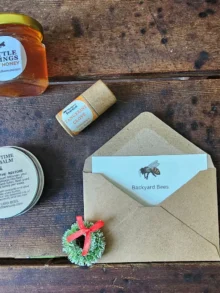 Backyard Bees holiday gift card (digital) styled with kraft envelope, evergreen wreath, raw honey, beeswax lip balm, and Sleepy Time Foot Balm on rustic wooden table