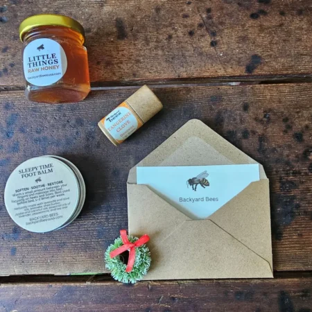 Backyard Bees holiday gift card (digital) styled with kraft envelope, evergreen wreath, raw honey, beeswax lip balm, and Sleepy Time Foot Balm on rustic wooden table