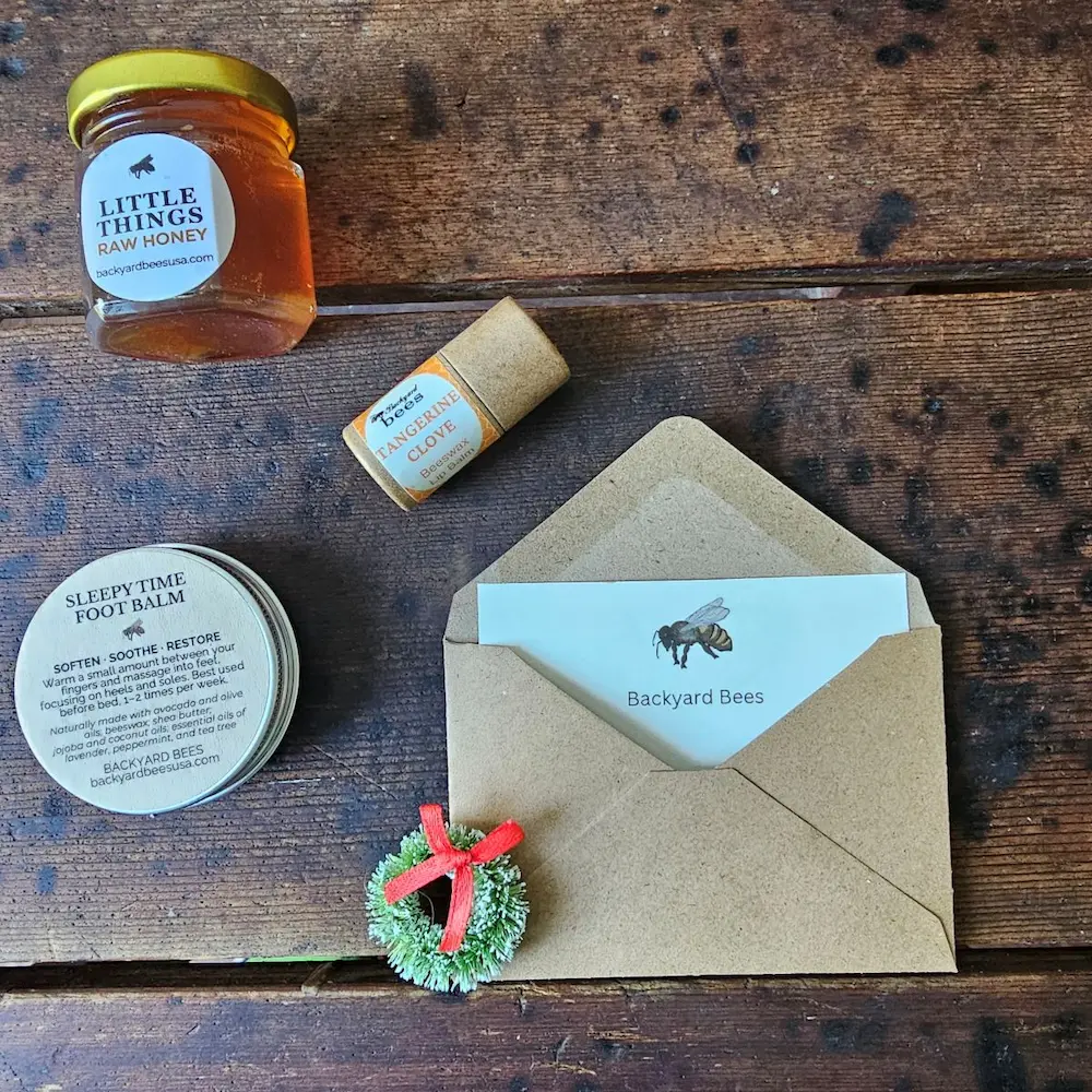 holiday gift card 3 Backyard Bees holiday gift card (digital) styled with kraft envelope, evergreen wreath, raw honey, beeswax lip balm, and Sleepy Time Foot Balm on rustic wooden table
