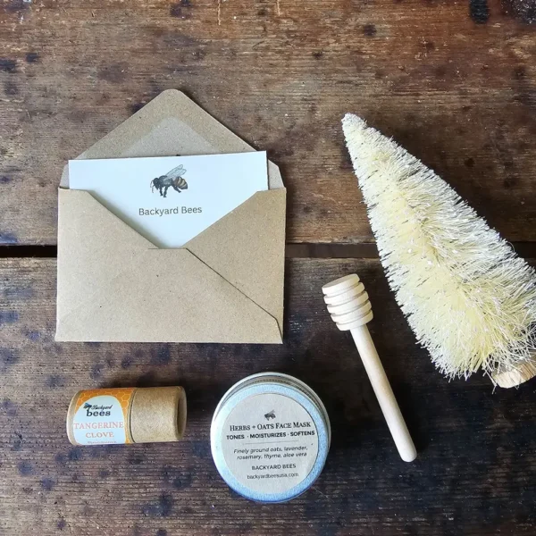 Backyard Bees holiday digital gift card styled with kraft envelope, honey dipper, white bottle brush tree, herbal face mask, and beeswax lip balm on rustic wooden table