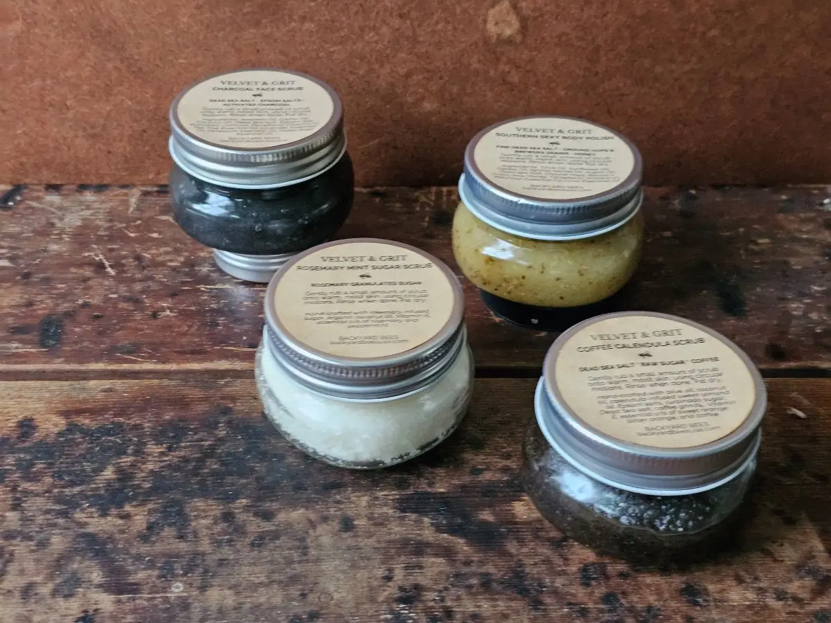 Four Velvet & Grit exfoliating body scrubs by Backyard Bees in glass jars on a rustic wooden background