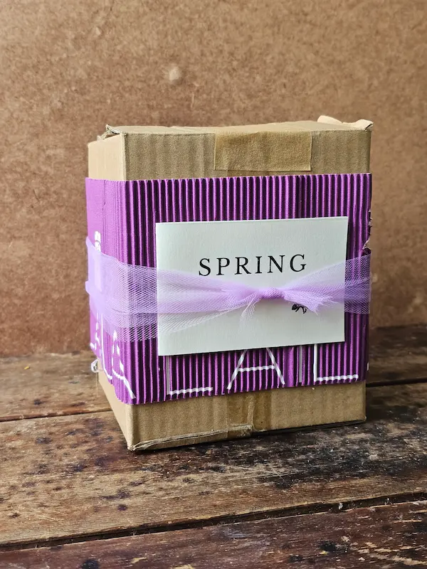 Closed Spring Basket gift box wrapped in purple corrugated cardboard with “Bloom” keepsake card and soft organza ribbon.